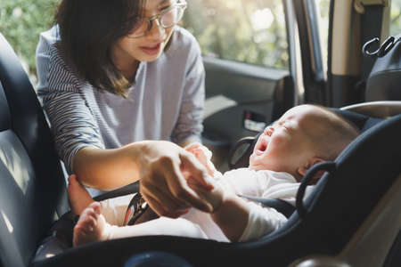 Beautiful Asian young mother comforting her baby boy crying while putting and fasten seat belts on his car seat in car.の写真素材