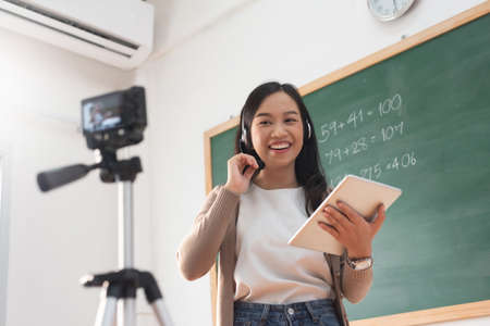 Young Asian woman teacher using digital camera standing online teaching with headphone and digital tablet in classroom at school.の写真素材