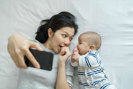 Young mother and newborn baby lying on bed taking selfie together with mobile phone at home. Asian Mom and son making video call with smartphoneの写真素材