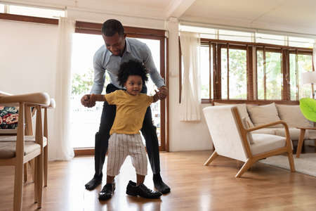 Smiling African little child having fun stepping with his dad shoe in living room at home. Happy small African American kid playing with father. Loving Black Family. Curly hair in afro style.の写真素材