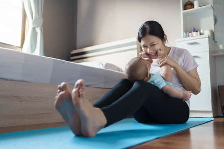 Happy smiling Asian young mother in sportswear playing and holding small baby son feet while...