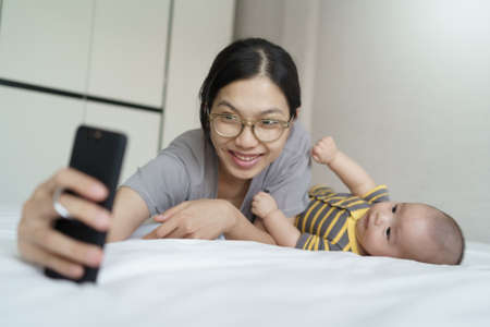 Happy Smiling Asian Young mother and newborn baby boy lying on bed taking selfie with mobile phone...