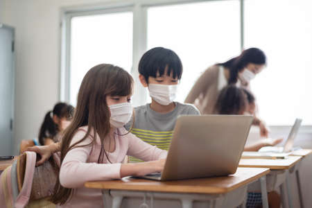 Diverse Multi Ethnic little girls and boys wearing protective face masks using laptop for studying in classroom at primary school. Education and Learning online technology.の写真素材