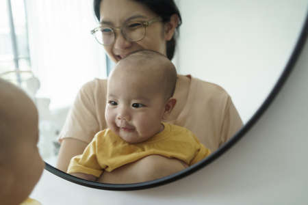 Playful Asian mother and little newborn baby boy having fun looking at their faces in mirror at...