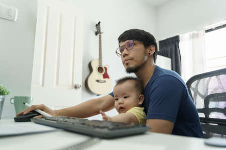 Asian Father working from home and taking care his little baby son. Designer male using computer during quarantine.の写真素材