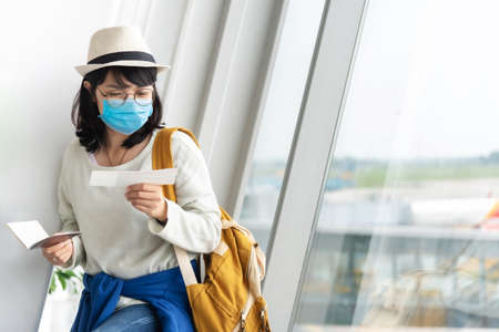 Asian Female tourist wearing protective face mask holding boarding pass waiting for the flight near the window of the airport terminal on her vacation. New normal Lifestyleの写真素材