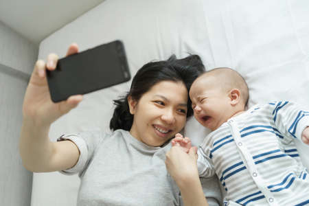 Asian Young Mother comforting or consoling her little toddler son while holding mobile phone. Sadness Newborn baby boy crying lying on bed.の写真素材