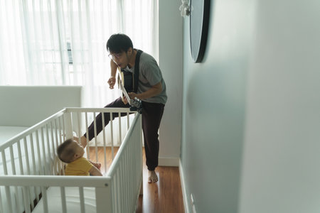 Happy Asian Dad singing and playing acoustic guitar while little adorable baby boy son sitting in the crib. Family having fun and enjoying in the music together. Hobbies and Leisure Lifestyle at home.の写真素材