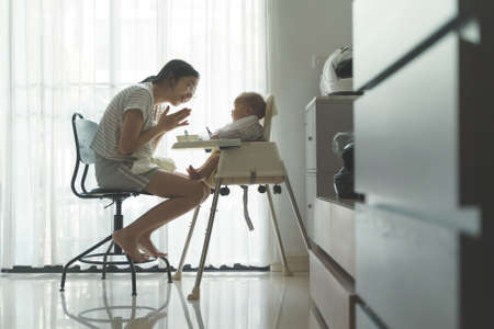 Happy Smiling Asian Young Mother enjoying feeding to her Little cute baby boy while sitting on high chair at home. Cheerful Family in breakfast or lunch time. Mom with first food of her adorable son.の写真素材