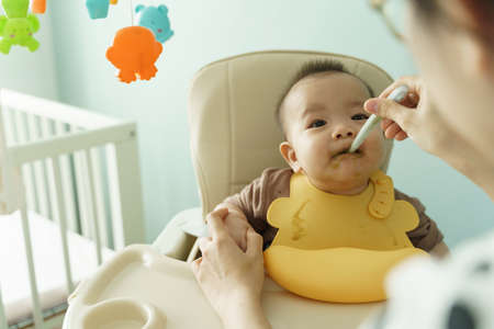 Asian Adorable little baby boy wearing yellow bib eating and sitting on high chair at home. Mother feeding pureed food to her son with a spoon.の写真素材