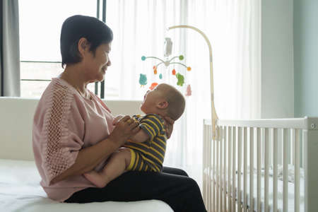 Happy Asian grandmother sitting on bed holding her little Grandchild in bedroom at home. Playful Adorable baby boy playing with Senior woman. Family having fun.の写真素材
