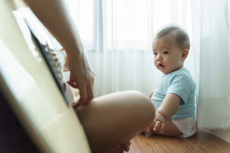 Asian little Cute boy sitting on wood floor looking his mother playing acoustic guitar. Happy Family enjoying on holiday. Hobbies and Leisure Lifestyle at home.の写真素材