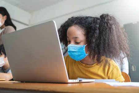 Adorable Black African American girl wearing protective face mask using laptop for studying in classroom at international school. Education and E-Learning with technologyの写真素材