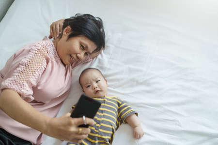 Happy Asian Grandmother and little baby boy lying on bed taking selfie photos with mobile phone. Senior woman and Grandchild or Grandson doing video call on smartphone with family togetherの写真素材