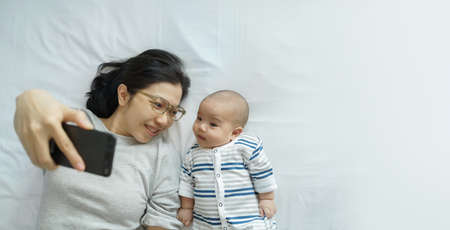 Young mother and little cute baby boy lying on bed taking selfie photos with mobile phone at home together. Asian Mom and son having video call on smartphone. Family, Modern Technology and people concept.の写真素材