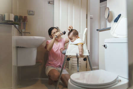 Asian Adorable little baby son sitting on high chair having haircut in the bathroom with hair clipper at home by his Mother.の写真素材