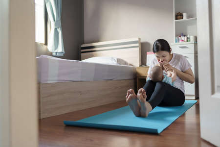Happy Asian young mother in playing and holding and kissing her newborn or infant baby feet while practicing yoga in bedroom at home.の写真素材