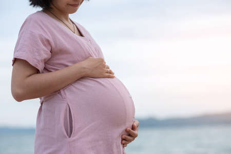 Happy Asian Pregnant woman touching her belly at the beach outdoor.の写真素材