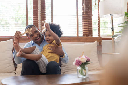 Cheerful African American Father and little son sitting on sofa playing wooden airplane toy at home together. Black family concept.の写真素材