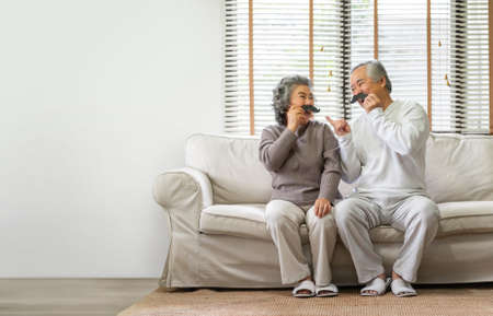 Happy Asian Senior Couple sitting on sofa and playing fake mustaches at home togetherness.の写真素材