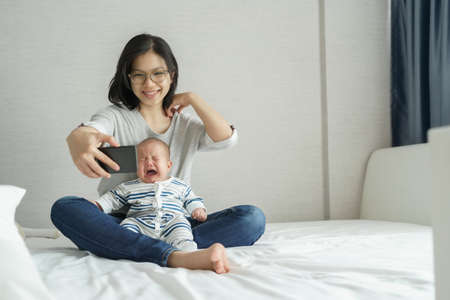 Smiling Asian Young Mother comforting or consoling her little baby son while doing online video call on smartphone. Sadness Newborn boy crying while taking selfie photo on mobile phone with Mom on bedの写真素材