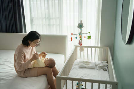 Asian  young mother playing with her little baby son in bedroom. Mom sitting on bed holding newborn child hands.の写真素材