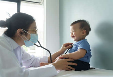 Asian female doctor wearing face mask examining little cute Baby boy with stethoscope in medical room at hospital.の写真素材