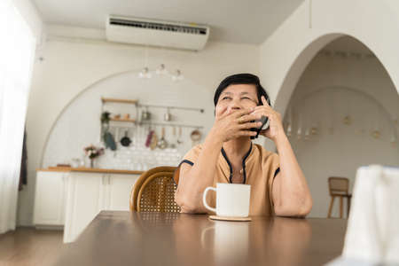 Happy Asian senior woman calling on smartphone sitting at table. Mature Female laughing and talking on her phone in kitchen at home.の写真素材