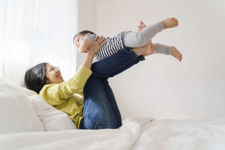 Loving Chinese Mom relax playing with her Adorable son at home, Happy Asian young mother lying on bed lifting little cute boy in the air.の写真素材