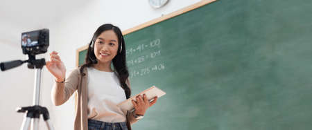Beautiful Asian woman teacher using digital camera and digital tablet standing online teaching with headphone and digital tablet in classroom at school, Banner, Panoramic, Web.の写真素材