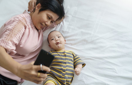Asian Grandmother and little baby boy lying on bed using smartphone at home.の写真素材