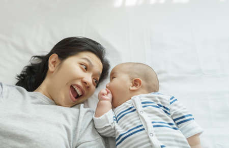 Loving Smiling Asian Young mother with her little cute baby son playing lying on bed together at...