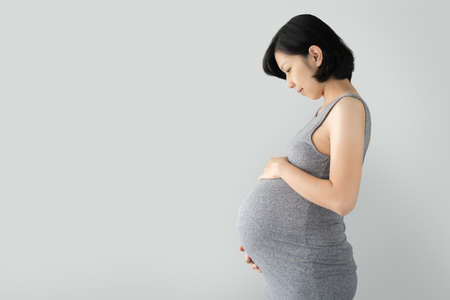 Pregnant Asian woman standing and touching big belly with her hands on gray Isolated background, Beautiful woman holding her stomachの写真素材