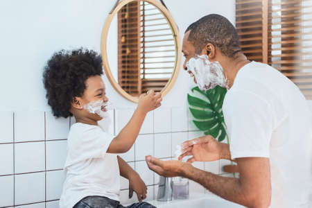 Loving African American Father and little boy having fun playing shaving foam on their faces in bathroom together, Happy African Dad and son.の写真素材