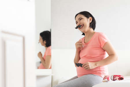 Portrait of Happy Asian pregnant woman playing paper moustaches pretending to be Father on her face anh tummy, Single Mom.の写真素材