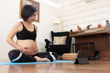 Asian pregnant woman practicing yoga online with digital tablet at home.の写真素材