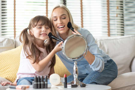 Attractive Happy Smiling Mother and little Daughter looking at the mirror and applying cosmetic with brushes on the cheekbones.の写真素材