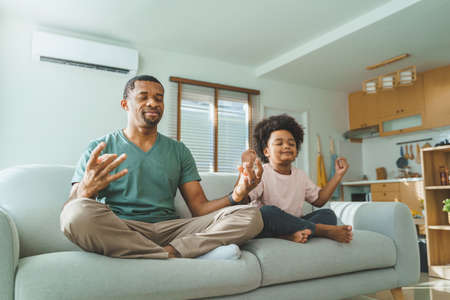 Happy Black African American father and Cute boy with eyes closed sitting on couch meditating at home together.の写真素材