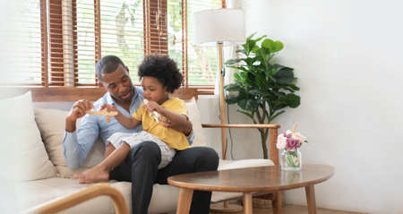 African American Father and little son sitting on couch playing wooden airplane toys in Living room at home together.の写真素材