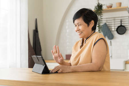 Happy Smiling Asian Mature adult woman having online video chat with friends or family on digital tablet at kitchenの写真素材