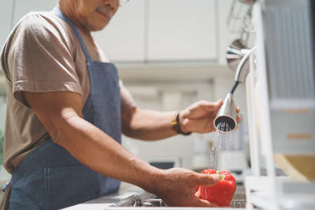 Selective focus on hands Asian healthy senior man preparing washing fresh red bell pepper or vegetables splashing in the kitchen sink at home.の写真素材