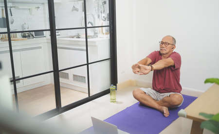 Asian Mature adult man wearing eyeglasses sitting on yoga mat stretching his arms after home exerciseの写真素材