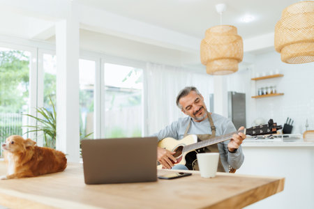 Attractive bearded Mature Adult Male wearing apron playing acoustic guitar, Using Laptop, Man with dog at homeの写真素材