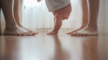 Close up feet of Family, Little baby learning to walk with Mother and Father on wooden floor at home. Cute toddler enjoying the first steps with parentの写真素材
