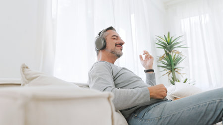 Attractive Bearded mature adult man sitting on the couch playing an air guitar and listening to music with headphones, Lifestyle concept, Relax in living room at home.の写真素材