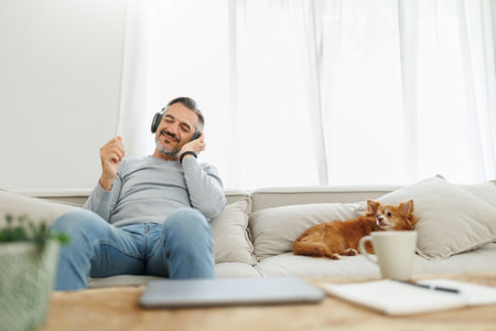 Attractive Bearded mature adult man sitting on the couch with his dog listening to music with headphones, Lifestyle concept, Relax in living room at home.の写真素材