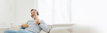 Bearded Senior mature adult man sitting on sofa listening to music with headphones and playing an air guitar, Lifestyle concept, Relaxation at home. Panoramic, Banner.の写真素材