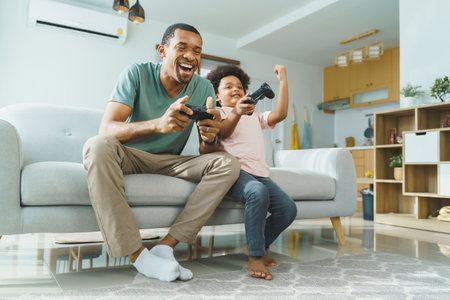 Cheerful Black African American Father and his Little son in Afro hair sitting on sofa using joysticks or game controllers playing console video games at homeの写真素材