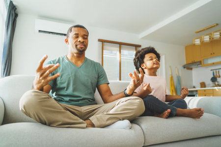 African American father and Cute boy smiling relaxed with closed eyes sitting on couch meditating together, Black Family practicing yoga at homeの写真素材