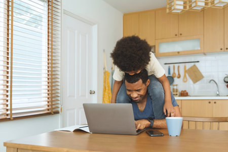 Cheerful Black African American Father playing with his little son while working at home, African Dad using laptop and taking care his boy at kitchenの写真素材
